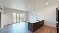 Located along Kruse Way in Lake Oswego, Oregon, this single-floor residence showcases a thoughtfully designed open-concept layout with contemporary finishes and high-quality materials throughout. The living and kitchen area form the centerpiece of the home, where light herringbone-patterned hardwood flooring extends across the space, complemented by crisp white walls and abundant natural light from large French doors that open directly onto a private balcony with tree-lined views.

The kitchen is well-appointed with dark shaker-style cabinetry featuring brass hardware, paired with upper white cabinetry for contrast. A spacious island with a white quartz countertop and undermount sink anchors the space, illuminated by globe pendant lights with brass accents. A gas range, built-in microwave, and subway tile backsplash complete the culinary space.

The primary bathroom offers a dual-sink vanity with a quartz countertop, dark shaker cabinetry, wall-mounted sconce lighting, and a walk-in shower. A secondary bathroom includes a tiled tub-shower combination and wood-look flooring. Additional features include a barn-style sliding door, a wall-mounted mini-split HVAC unit, and a herringbone-floored living area with balcony access, creating a cohesive and modern living environment.