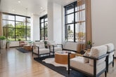 The lobby at Jules on 3rd in Boise, Idaho is a thoughtfully designed, generously proportioned space that blends contemporary style with welcoming functionality. Striking herringbone-patterned hardwood floors run throughout, tying together the various zones within the open layout. Soaring floor-to-ceiling windows flood the interior with abundant natural light while offering views of the surrounding streetscape and greenery.

The main entrance features a dramatic glass curtain wall framed in matte black, creating a bold architectural statement. A central reception desk anchored by elegant pendant lights serves as the heart of the lobby, flanked by comfortable white lounge seating arranged around a warm wood coffee table. Nearby, a dedicated lounge area showcases modern accent chairs, a shimmering decorative wall installation, and a wall-mounted display screen.

A dining area with high-top and standard seating lines the window wall, ideal for casual work or socializing. A private meeting room offers a round dark-wood table beneath a dramatic black-and-gold dome pendant, textured wallcovering, and a wall-mounted screen. An additional lounge nook features nature-inspired wallpaper, bar stools, and natural light filtering through sheer curtains. The hallways connect each zone seamlessly, maintaining the space's cohesive, upscale aesthetic throughout.
