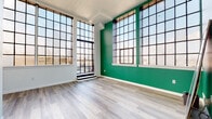 This single-floor space blends industrial character with modern finishes throughout its well-appointed layout. The most striking feature is the main living area, where oversized, multi-pane steel-framed warehouse windows stretch toward the ceiling on two walls, flooding the room with abundant natural light and offering open views of the surrounding cityscape. A bold emerald green accent wall anchors this space, complementing the light grey wood-style flooring that runs consistently throughout the unit.

The kitchen is outfitted with white cabinetry, dark stone countertops featuring subtle veining, a stainless steel sink, an electric range, and a classic white subway tile backsplash, delivering a clean, functional aesthetic.

The bedrooms each carry their own character through vivid accent walls — one featuring a vibrant cobalt blue — paired with the same light wood flooring. One bedroom includes a built-in closet with shelving, while another benefits from its own industrial-style multi-pane window, maintaining the cohesive design language of the space. Additional rooms present neutral white walls and ample natural light. Throughout, white baseboards and clean lines contribute to a bright, contemporary ambiance.