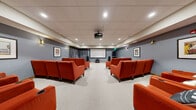 The Theater Room at 500 Apex Dr in Windsor, Colorado, is a thoughtfully designed private screening space that delivers an immersive cinematic experience. The room features a ceiling-mounted projector paired with a large projection screen at one end, flanked by floor-to-ceiling gray drape panels that enhance the theatrical atmosphere. Recessed lighting and wall-mounted sconces provide warm, adjustable illumination throughout the space.

Seating consists of numerous bold, rust-orange upholstered armchairs arranged in rows across the carpeted floor, each accented with movie-themed throw pillows. The chairs are generously spaced, allowing for comfortable viewing from every angle. The soft, light-colored carpet and cool blue-gray walls create a polished, inviting ambiance.

At the rear of the room, a built-in kitchenette with dark wood cabinetry, under-counter refrigeration, and under-cabinet lighting provides convenient refreshment preparation. A classic popcorn machine sits atop the counter, adding an authentic movie-night touch. Framed cinema-themed artwork adorns the walls, reinforcing the room's dedicated purpose. The space is well-suited for group screenings, presentations, or community entertainment gatherings.