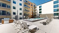 This contemporary multi-story residential complex is designed with a striking exterior that combines warm wood-tone cladding, neutral stucco panels, and large black-framed windows. Numerous private balconies with sleek metal railings line the building's façade across multiple floors, offering elevated outdoor space for residents.

The centerpiece of the property is a well-appointed outdoor amenity deck spanning two levels. A generously sized rectangular swimming pool with tiled surrounds anchors the space, complemented by a separate spa. The pool area is equipped with an accessibility lift and lined with rows of white lounge chairs, ideal for sunbathing beneath open skies. Black metal perimeter fencing provides both security and a clean architectural edge throughout the deck.

Adjacent to the pool, a spacious courtyard lounge features large-format concrete pavers, cylindrical planter seating elements, and overhead string lights that create an inviting atmosphere for evening gatherings. The open-air design of the courtyard is framed by the building's interior-facing facades, creating a sheltered yet airy environment.

The walkways connecting the amenity areas feature sturdy metal railing systems and broad pathways, ensuring ease of movement throughout. The overall design reflects a modern urban aesthetic, balancing functionality with contemporary style.