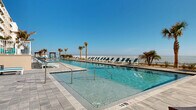 Located directly on North Atlantic Avenue in Daytona Beach, Florida, this outdoor amenity level delivers an exceptional beachfront experience. The centerpiece is a generously sized swimming pool with crystal-clear turquoise water, framed by a broad, paver-tiled sun deck designed to maximize relaxation and enjoyment. Rows of teal-cushioned lounge chairs and white-framed chaise lounges are thoughtfully arranged throughout the deck, providing ample space for sunbathing and leisure.

Tall palm trees are strategically placed across the pool area, offering both tropical ambiance and natural shade, while a pergola-style structure provides an additional shaded retreat. The pool deck is oriented to face the Atlantic Ocean directly, with a white-railed perimeter fence allowing unobstructed views of the shoreline and open sky beyond.

A wide, paved walkway runs alongside the multi-story building, connecting various sections of the outdoor space and flanked by low-maintenance coastal landscaping featuring ornamental grasses, native plantings, and decorative stone. Stairways with white railings provide access between levels of the building. The overall design reflects a clean, modern coastal aesthetic, with the building's neutral facade and large balconies complementing the bright, sun-drenched atmosphere of this premier beachside outdoor space.
