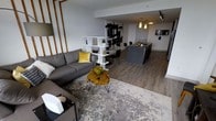 This single-floor space showcases a thoughtfully designed open-concept layout that seamlessly connects the living, dining, and kitchen areas into one cohesive environment. The living area is anchored by a plush grey sectional sofa paired with a striking live-edge wooden coffee table on a textured area rug, lending a warm, organic contrast to the otherwise contemporary aesthetic. A white geometric open-shelf unit serves as a subtle room divider, defining the living space while maintaining visual openness throughout. Warm golden accents, including throw pillows and decorative elements, complement the neutral grey and white palette. The kitchen features a sleek island with a dark countertop and pendant lighting overhead, offering both a functional prep space and casual seating. Wood-toned vertical wall paneling in the living area adds architectural interest and warmth. Light wood-style laminate flooring runs throughout, tying the spaces together cohesively. The overall ambiance is modern and curated, balancing clean lines with natural textures for a welcoming, sophisticated feel.