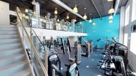 This two-floor fitness facility is designed to accommodate a wide range of training needs within a spacious, well-equipped environment. The main floor is lined with an extensive selection of strength machines, cable systems, and cardio equipment, all arranged to maximize floor space and ease of movement between stations. The flooring throughout is a durable, dark rubberized surface suited for high-traffic gym use.

A defining architectural feature is the open mezzanine level, accessible via a staircase with sleek glass and metal railings that lend a modern, industrial aesthetic to the space. The upper level mirrors the ground floor with additional strength equipment, providing ample capacity for members at peak times.

Visually, the facility is anchored by bold turquoise accent walls that energize the atmosphere and give the space a vibrant, contemporary identity. Overhead, exposed ductwork and dark ceiling panels reinforce the industrial design language, while pendant-style pendant lights are suspended throughout both levels, ensuring consistent, warm illumination across the entire facility. The combination of open sightlines between floors, high ceilings, and thoughtful equipment placement creates an expansive and motivating training environment suited for fitness enthusiasts of all levels.