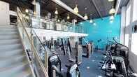This two-floor fitness facility is designed to accommodate a wide range of training needs within a spacious, well-equipped environment. The main floor is lined with an extensive selection of strength machines, cable systems, and cardio equipment, all arranged to maximize floor space and ease of movement between stations. The flooring throughout is a durable, dark rubberized surface suited for high-traffic gym use.

A defining architectural feature is the open mezzanine level, accessible via a staircase with sleek glass and metal railings that lend a modern, industrial aesthetic to the space. The upper level mirrors the ground floor with additional strength equipment, providing ample capacity for members at peak times.

Visually, the facility is anchored by bold turquoise accent walls that energize the atmosphere and give the space a vibrant, contemporary identity. Overhead, exposed ductwork and dark ceiling panels reinforce the industrial design language, while pendant-style pendant lights are suspended throughout both levels, ensuring consistent, warm illumination across the entire facility. The combination of open sightlines between floors, high ceilings, and thoughtful equipment placement creates an expansive and motivating training environment suited for fitness enthusiasts of all levels.