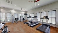 This fitness center offers a comprehensive workout environment within a single, open-plan space. The room is finished with warm-toned herringbone-patterned carpet tiles that lend a polished, modern aesthetic to the floor throughout. Crisp white walls and crown molding contribute to a clean, well-maintained appearance, while recessed lighting and multiple ceiling fans ensure the space remains comfortable and well-lit during use.

A generous arrangement of windows lines the perimeter walls, fitted with horizontal blinds that allow natural light to filter in while maintaining privacy. The abundant daylighting creates a bright, energizing atmosphere well-suited to physical activity.

The cardio zone features multiple treadmills and elliptical trainers from recognized brands including True and Precor, arranged to make efficient use of the available floor area. The strength training section includes Hoist selectorized machines, a multi-function cable system, a free weight rack with dumbbells, and a weight bench. A large framed wall mirror is positioned to allow users to monitor their form during exercises.

Wall-mounted flat-screen televisions are placed at strategic viewing angles throughout the room. The space also includes a sanitation station for equipment hygiene. Overall, the fitness center provides a fully functional and thoughtfully organized facility suitable for a wide range of workout routines.