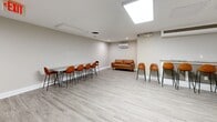 Located at 1131 W University Blvd in Silver Spring, Maryland, this single-floor space offers a clean, modern layout that seamlessly integrates a kitchen and living area into one cohesive environment. The open-concept design maximizes functionality while maintaining a welcoming atmosphere throughout.

The living area is bright and well-appointed, featuring light gray luxury vinyl plank flooring that runs throughout the entire space. A skylight overhead draws in natural light, complementing the recessed lighting and flush-mounted fixtures in the ceiling. A wall-mounted mini-split HVAC unit ensures year-round climate comfort. The seating arrangement includes a warm cognac leather sofa and multiple leather bar-height chairs paired with long high tables, making the space well-suited for both casual lounging and collaborative use.

The kitchen is thoughtfully designed with dark espresso cabinetry, stainless steel appliances including a full-size refrigerator, microwave, and dishwasher, and granite-style countertops. A mosaic tile backsplash in earthy tones adds a decorative touch. A secondary mini-split unit in the kitchen area provides dedicated climate control. The neutral gray walls throughout create a clean, professional backdrop that ties the two zones together cohesively.
