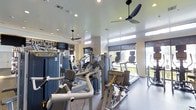 This single-level fitness center offers a comprehensive workout environment designed to accommodate a variety of exercise routines. The space is outfitted with an extensive selection of equipment, including treadmills, ellipticals, and multiple cable and resistance machines arranged to maximize usability and flow throughout the room.

Natural light filters in through a series of clerestory windows positioned along the upper walls, as well as larger floor-to-ceiling windows and a glass door that open to an outdoor view, creating a bright and energizing atmosphere. Roller shades provide the option to control light levels as needed. Recessed ceiling lighting supplements natural light, ensuring the space remains well-lit at all times.

Two modern ceiling fans are mounted overhead, promoting air circulation throughout the facility. The flooring features a durable, speckled surface suited for high-traffic athletic use. A decorative typographic mural with urban-themed text adds a local character to one wall, accompanied by mounted flat-screen televisions for entertainment during workouts. The overall design balances functionality with a clean, contemporary aesthetic.
