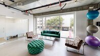 Located at 616 S Kenmore Ave in Los Angeles, this combined gym and recreation room offers a well-equipped, industrial-modern facility across a single floor. The space is defined by its exposed concrete ceilings, visible ductwork, and expansive floor-to-ceiling windows that flood the interior with natural light and provide open views of the surrounding streetscape and tree canopy.

The fitness area is divided into distinct zones. The cardio section features multiple treadmills and stationary bikes positioned directly in front of the large windows, with a wall-mounted flat-screen TV overhead for entertainment during workouts. A dedicated free weights area includes a full rack of dumbbells, an adjustable workout bench, resistance balls, and foam rollers, with mirrored walls enhancing the sense of space. Rubber flooring throughout the workout zones provides safety and durability.

Adjacent to the gym is a recreation and lounge area furnished with tufted velvet seating — including a bold green sofa, coordinating armchairs, and an ottoman — arranged around a glass coffee table. This area benefits from the same expansive window views, creating a bright and inviting atmosphere for relaxation.

A kitchenette serves the space with modern cabinetry, a stainless steel sink, a pod coffee machine, microwave, under-counter beverage refrigerator, and a wall-mounted TV, making it a convenient amenity hub for residents.