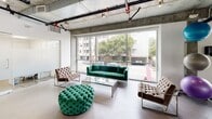 Located at 616 S Kenmore Ave in Los Angeles, this combined gym and recreation room offers a well-equipped, industrial-modern facility across a single floor. The space is defined by its exposed concrete ceilings, visible ductwork, and expansive floor-to-ceiling windows that flood the interior with natural light and provide open views of the surrounding streetscape and tree canopy.

The fitness area is divided into distinct zones. The cardio section features multiple treadmills and stationary bikes positioned directly in front of the large windows, with a wall-mounted flat-screen TV overhead for entertainment during workouts. A dedicated free weights area includes a full rack of dumbbells, an adjustable workout bench, resistance balls, and foam rollers, with mirrored walls enhancing the sense of space. Rubber flooring throughout the workout zones provides safety and durability.

Adjacent to the gym is a recreation and lounge area furnished with tufted velvet seating — including a bold green sofa, coordinating armchairs, and an ottoman — arranged around a glass coffee table. This area benefits from the same expansive window views, creating a bright and inviting atmosphere for relaxation.

A kitchenette serves the space with modern cabinetry, a stainless steel sink, a pod coffee machine, microwave, under-counter beverage refrigerator, and a wall-mounted TV, making it a convenient amenity hub for residents.