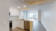This single-floor space showcases a thoughtfully designed open-concept layout that seamlessly connects the kitchen and living areas. The kitchen is outfitted with crisp white cabinetry, a classic white subway tile backsplash, and light granite countertops that lend a clean, polished aesthetic. A peninsula with a stainless steel dishwasher and an undermount sink with a modern brushed-nickel faucet provides both functionality and a natural division between the kitchen and the main living area. The flooring throughout features warm-toned hardwood-style planks that contrast beautifully with the bright white walls and cabinetry. Recessed lighting in the kitchen ensures ample illumination, while the open living area benefits from an abundance of natural light streaming through expansive floor-to-ceiling windows. These large windows frame impressive views of the surrounding urban skyline, filling the space with a sense of openness and connection to the city beyond. The living area's neutral palette and minimalist design create a versatile canvas ready for personalization. The overall ambiance is modern, airy, and sophisticated, with a cohesive design language that balances style and practicality throughout the space.