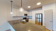 This single-floor unit offers a clean, modern aesthetic with a thoughtful layout visible in the open floor plan. The kitchen serves as a central highlight, showcasing white flat-front cabinetry extending to the ceiling, a classic subway tile backsplash, and dark quartz countertops. Stainless steel appliances, including an electric range, built-in microwave, and a full-size refrigerator, complement the space. A large kitchen island with an integrated sink and pendant globe lighting above adds both functionality and style, while recessed lighting ensures the area remains well-lit.

The living area features light wood-style flooring, a ceiling fan, and multiple windows that allow natural light to fill the room. The bedroom is finished with plush carpeting, a ceiling fan, and window access. A spacious walk-in closet is equipped with wire shelving and hanging rods along multiple walls, offering generous storage capacity.

The bathroom includes wood-style flooring, a tub-shower combination with white tile surround, white cabinetry with dark countertops, and wall-mounted lighting above the vanity mirror. Throughout the unit, white walls and clean finishes create a bright, contemporary atmosphere.