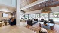 This single-floor clubhouse space is defined by its striking architectural character and thoughtfully curated interiors. The main lounge area commands attention with soaring vaulted ceilings clad in warm, reclaimed-style wood planking, creating a rich contrast against the crisp white walls. Large floor-to-ceiling windows line the exterior wall, flooding the space with natural light and offering unobstructed views of the outdoor pool and tree-lined grounds beyond.

The lounge is furnished with a mix of leather sofas, wood-framed accent chairs, and layered area rugs in Southwestern-inspired patterns, establishing a comfortable yet refined atmosphere. Oversized woven pendant light fixtures add an organic, artisanal touch overhead. A secondary seating area provides additional casual gathering space, anchored by a sculptural stone coffee table and complementary accent furniture.

The kitchen area features a large dark stone-topped island with matte black pendant lighting, warm wood cabinetry, and a patterned kilim runner underfoot. Adjacent to the kitchen, a cozy dining nook with tufted leather banquette seating and bistro-style tables overlooks the fitness center through an interior window. Light-toned tile flooring flows consistently throughout, tying all areas together into one cohesive, welcoming community space.