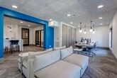 Located at 13220 Foster St in Overland Park, Kansas, this single-floor amenity space is thoughtfully designed to support both productivity and relaxation. The interior is defined by a clean, contemporary aesthetic, with crisp white walls accented by a bold teal feature wall that serves as an eye-catching architectural element throughout the space.

The open coworking area is equipped with long shared desks, multiple computer workstations, and ergonomic seating, all illuminated by a combination of stylish pendant lighting — including globe and geometric fixtures — and recessed ceiling lights. Herringbone-patterned carpet tile flooring runs throughout, adding texture and visual interest underfoot, while a concrete-finish ceiling lends an industrial yet refined character to the overall design.

A glass-walled conference room with black metal framing provides a semi-private meeting space, offering transparency while maintaining a sense of enclosure. Adjacent lounge seating, including a large, plush sectional sofa, creates a comfortable area for informal gatherings or relaxed work sessions. A bar-height counter with seating is visible nearby, suggesting a casual dining or social nook.

Natural light filters in through multiple large windows, connecting the interior to the surrounding exterior. The layout flows openly between zones, making this a versatile, well-appointed space suited for community use.