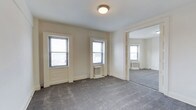 This single-floor space presents a straightforward, functional layout characterized by neutral cream-colored walls and gray wall-to-wall carpeting that extends throughout the connected rooms. The main room is illuminated by two double-hung windows fitted with blinds, allowing natural light to filter in, while a flush-mount ceiling light fixture provides overhead lighting. Two compact radiator units are positioned beneath the windows, indicating a traditional steam or hot water heating system — a hallmark of classic building construction. White-painted door and window trim adds a clean contrast against the neutral walls. An open doorway connects the main room to an adjoining room, which mirrors the design with its own radiator unit and window, maintaining a consistent aesthetic throughout the space. The layout suggests a straightforward flow between rooms, suitable for a variety of uses. The overall ambiance is understated and neutral, offering a blank canvas that can be adapted to different needs. The architectural details, including the classic window style and radiator placement, point to a building of traditional construction and character.