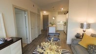 This single-floor residence features an open-concept layout that connects the living, dining, and kitchen areas into one cohesive, functional space. Dark hardwood flooring runs throughout, providing a warm contrast to the bright white walls and cabinetry. The kitchen is positioned toward the rear of the open floor plan and is equipped with white shaker-style cabinetry, stainless steel appliances, and a central island with a butcher block countertop that doubles as a casual dining or prep area. Pendant lighting above the island and track lighting on the ceiling provide targeted illumination throughout the kitchen zone. The living area at the front of the space is accented by a striped area rug and modern accent chairs, adding visual interest. Multiple interior doors along the left wall suggest access to additional rooms such as bedrooms or bathrooms, offering a degree of privacy within the unit. The front entry door, finished in a bold gray tone, serves as a subtle focal point within the layout. Overall, the space presents a clean, contemporary aesthetic with practical design choices throughout.