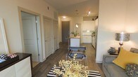 This single-floor residence features an open-concept layout that connects the living, dining, and kitchen areas into one cohesive, functional space. Dark hardwood flooring runs throughout, providing a warm contrast to the bright white walls and cabinetry. The kitchen is positioned toward the rear of the open floor plan and is equipped with white shaker-style cabinetry, stainless steel appliances, and a central island with a butcher block countertop that doubles as a casual dining or prep area. Pendant lighting above the island and track lighting on the ceiling provide targeted illumination throughout the kitchen zone. The living area at the front of the space is accented by a striped area rug and modern accent chairs, adding visual interest. Multiple interior doors along the left wall suggest access to additional rooms such as bedrooms or bathrooms, offering a degree of privacy within the unit. The front entry door, finished in a bold gray tone, serves as a subtle focal point within the layout. Overall, the space presents a clean, contemporary aesthetic with practical design choices throughout.