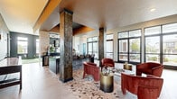 This well-appointed amenity space spans a generous single floor, thoughtfully designed to offer both social and recreational functionality. The main lounge area makes an immediate impression with its striking terrazzo flooring, featuring an eclectic mosaic of earthy tones that anchors the room. Towering stone-clad columns rise to meet warm gold-toned ceilings, adding architectural drama and a sense of vertical scale. Plush velvet lounge chairs in a rich terracotta hue are arranged around sleek metallic accent tables, creating inviting conversation clusters. Globe pendant lights in brushed gold punctuate the deep navy accent walls, lending a sophisticated, curated ambiance. A bar counter with bar-height seating runs along one side of the lounge, ideal for casual gatherings. Floor-to-ceiling windows and glass doors line multiple walls throughout, flooding the space with natural light and offering open views of the surrounding exterior. Adjacent to the lounge is a dedicated indoor putting green, complete with regulation-style turf and flags, framed by a white brick accent wall with a mounted flat-screen television and stylish wall sconces. A comfortable seating area nearby makes this zone a versatile recreational retreat. The overall design blends modern elegance with welcoming warmth.