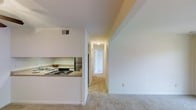 This single-floor residential unit offers a clean, functional layout designed for comfortable everyday living. The open-concept design seamlessly connects the kitchen to the main living area, creating a sense of spaciousness throughout. The kitchen features a dark electric cooktop, a stainless steel refrigerator, and a countertop with ample workspace, all set against bright white cabinetry. A peninsula-style counter provides a natural division between the kitchen and the adjoining living space, which is finished with plush wall-to-wall carpeting in a warm neutral tone. Crisp white walls throughout the unit contribute to a bright and airy ambiance. A hallway extends from the living area, leading to additional rooms including what appears to be a bedroom and closet space, suggesting a practical and well-organized floor plan. Ceiling-mounted lighting fixtures provide consistent illumination throughout the hallway and living spaces. A ceiling vent ensures proper climate control, adding to the overall comfort of the unit. The overall aesthetic is simple, clean, and move-in ready, making it an accessible and straightforward living space.