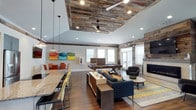 This single-floor community space masterfully blends rustic charm with modern design. The centerpiece of the room is a striking reclaimed wood ceiling inset, framed by clean white drywall and complemented by matching wood-paneled accent walls that lend warmth and texture throughout the space. Twin ceiling fans and recessed lighting ensure the area remains bright and comfortable at all times.

The open-plan layout is thoughtfully zoned for a variety of activities. A large lounge area anchors the space with a sleek sectional sofa, accent chairs, and a central coffee table arranged atop a bold area rug, creating an inviting setting for conversation and relaxation. Adjacent to this is a pool table, offering a dedicated games zone. A built-in fireplace with a stone surround adds a cozy focal point on one wall, while multiple windows allow natural light to filter throughout the room.

A granite-topped bar counter with bar seating provides a social hub, ideal for casual dining or gathering, while a separate dining area with vibrant orange chairs adds a pop of color. Hardwood flooring ties the entire space together, contributing to the warm, welcoming ambiance. The overall aesthetic is a harmonious mix of industrial, rustic, and contemporary elements.