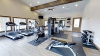 This fitness center combines rustic charm with modern functionality, featuring a vaulted ceiling accented by an exposed wooden beam that adds warmth and character to the space. Recessed lighting and a ceiling fan ensure the room stays well-lit and comfortable during workouts. Light wood-look flooring runs throughout, complementing the neutral wall tones and contributing to a clean, inviting atmosphere.

The facility is outfitted with a solid selection of cardio equipment, including treadmills, an elliptical, and a stationary bike, each positioned on protective floor mats. A multi-station cable machine serves as the centerpiece for strength training, while an adjustable weight bench provides additional versatility for resistance exercises. A large wall mirror expands the sense of space and allows for form checks during workouts.

Natural light filters in through multiple windows along the walls, keeping the environment bright and energizing. Wall-mounted televisions offer entertainment options during exercise sessions. The open layout provides ample room to move freely between equipment, making the fitness center both practical and comfortable for a variety of training routines.