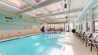 Located at 230 N 6th St in Richmond, Virginia, this indoor aquatic facility spans a generous single-floor footprint, offering a well-appointed and inviting environment for recreation and relaxation. The centerpiece of the space is a large, pristine indoor swimming pool with clearly marked depth indicators and pool entry railings, surrounded by a wide pool deck finished in light-toned flooring. Soft aqua-green walls, accented by white tile wainscoting, lend the space a clean and refreshing aesthetic throughout.

Overhead, a coffered skylight ceiling allows abundant natural light to flood the pool area, creating a bright and airy atmosphere even while fully enclosed. Wall-mounted sconce lighting supplements the natural light, ensuring the space remains well-lit at all times. Rows of sleek lounge chairs line the poolside, offering comfortable spots for relaxation before or after a swim, while additional seating areas with tables and chairs are positioned near the pool deck perimeter.

A standout feature is the expansive floor-to-ceiling glass wall that opens the space visually to an interior courtyard, filled with lush tropical plants and outdoor seating, further enhancing the sense of openness and connection to the surrounding building. Towel stations and utility storage are conveniently placed near pool access points, rounding out this thoughtfully designed aquatic retreat.
