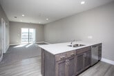 This well-appointed single-floor residence offers a clean, contemporary design throughout. The open-concept layout seamlessly connects the kitchen and living area, creating a spacious and functional common space. The kitchen is equipped with dark walnut-toned cabinetry, stainless steel appliances including a refrigerator, microwave, and range, and a large peninsula featuring a white quartz countertop with a double undermount sink — ideal for both meal preparation and casual dining. Light hardwood-style flooring runs through the kitchen and entry areas, transitioning to plush carpet in the living room and bedroom. The living area is bright and airy, benefiting from natural light that pours in through a sliding glass door leading to a private balcony with open views. The bedroom is generously sized, featuring a ceiling fan, a large window dressed with vertical blinds, and comfortable carpeted flooring. The full bathroom is finished with a dark wood vanity, white quartz countertop, a framed mirror with overhead lighting, and chrome fixtures. A walk-in closet is accessible from the bedroom area, providing ample storage. Neutral warm-gray walls and recessed lighting throughout contribute to the clean, modern ambiance of this thoughtfully designed residence.
