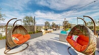 Located at 7650 Van Nuys Blvd in the Van Nuys neighborhood of Los Angeles, this expansive rooftop terrace offers a thoughtfully designed outdoor retreat with a lively, resort-inspired atmosphere. The open-air deck is finished with a smooth concrete surface and is generously appointed with multiple distinct seating zones, each suited to different social or relaxation needs.

Colorful woven hanging egg chairs with vibrant orange cushions anchor the space near the entrance, adding a playful and modern design element. A central lounge area features teak-framed sofas and chairs arranged around a bold, round fire pit bowl, creating an inviting focal point for gatherings. String lights are strung overhead throughout the terrace, lending a warm ambiance during evening hours.

A private cabana with a canopied daybed provides a shaded retreat, ideal for relaxing while taking in the panoramic views of the San Fernando Valley and the surrounding mountain ranges. Vivid turquoise planters filled with ornamental grasses and mature trees add lush greenery and color contrast across the deck.

A dedicated BBQ and dining area with outdoor grills and bar-height seating rounds out the amenities. The terrace overlooks the Van Nuys cityscape and distant mountain ridgelines, offering an open and airy setting well above the surrounding streetscape.