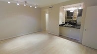 This single-floor space features an open-concept layout that seamlessly connects the living area to the kitchen. The living area is finished with light-toned hardwood flooring that runs throughout, creating a clean and cohesive aesthetic. A modern track lighting system is mounted on the ceiling, providing adjustable illumination across the main living space. The neutral, warm-beige walls contribute to a bright and inviting atmosphere.

The kitchen is positioned in an open pass-through configuration, offering both functionality and visual connection to the living area. It is equipped with dark cabinetry contrasted by bold black countertops and includes stainless steel appliances, among them a microwave and range. A stainless steel sink is integrated into the countertop, and pendant lighting above the kitchen area adds a refined touch. The kitchen's efficient layout maximizes workspace while maintaining an open feel.

A doorway visible from the main living area leads to additional spaces within the unit. Central air ventilation is integrated into the ceiling, ensuring climate comfort. Overall, the space presents a modern, minimalist character with practical finishes suited for everyday living.