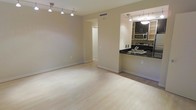 This single-floor space features an open-concept layout that seamlessly connects the living area to the kitchen. The living area is finished with light-toned hardwood flooring that runs throughout, creating a clean and cohesive aesthetic. A modern track lighting system is mounted on the ceiling, providing adjustable illumination across the main living space. The neutral, warm-beige walls contribute to a bright and inviting atmosphere.

The kitchen is positioned in an open pass-through configuration, offering both functionality and visual connection to the living area. It is equipped with dark cabinetry contrasted by bold black countertops and includes stainless steel appliances, among them a microwave and range. A stainless steel sink is integrated into the countertop, and pendant lighting above the kitchen area adds a refined touch. The kitchen's efficient layout maximizes workspace while maintaining an open feel.

A doorway visible from the main living area leads to additional spaces within the unit. Central air ventilation is integrated into the ceiling, ensuring climate comfort. Overall, the space presents a modern, minimalist character with practical finishes suited for everyday living.