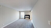 This single-floor unit offers a well-organized layout across several distinct living areas. The living room serves as the central gathering space, featuring plush gray carpeting, neutral gray walls, a textured ceiling, and a fireplace with a contrasting black surround and white mantel. A window provides natural light, while a sliding glass door fitted with vertical blinds opens to an outdoor view of a landscaped communal area.

The galley-style kitchen is efficiently designed with warm oak cabinetry lining both sides, granite-look countertops, a double stainless steel sink, and white appliances including an electric range and refrigerator. Light ceramic tile flooring runs throughout, and a dedicated pantry closet adds practical storage. The kitchen connects openly to a dining or transitional area, creating a functional flow.

The bedrooms are carpeted and finished in soft neutral tones, each offering natural light through windows and ample wall space. Closet access is available within the bedroom areas. The bathroom features a full tub-shower combination with clean white square tile surround. Throughout the unit, white baseboards and consistent neutral paint create a cohesive, understated aesthetic suited for a range of furnishing styles.