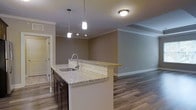 This single-floor space is designed with an open-concept layout that seamlessly connects the kitchen, dining, and living areas, creating a cohesive and functional flow throughout. The kitchen serves as a central focal point, featuring dark cabinetry, granite countertops with subtle veining, and a peninsula island equipped with an undermount sink and pendant lighting above. A subway tile backsplash adds a clean, modern touch, while stainless steel appliances complete the space. The peninsula extends into a raised bar-height surface, offering additional seating and naturally defining the transition between the kitchen and the adjoining living area.

Hardwood-style flooring runs throughout the main living spaces, lending warmth and visual continuity. Crown molding frames the ceilings, adding an elevated architectural detail. The living area is generously proportioned, offering flexible space for furniture arrangement. A separate room off the main living space provides additional privacy and versatility. Neutral-toned walls in warm gray complement the overall palette, creating a balanced and welcoming ambiance. The unit's thoughtful layout and quality finishes make it a polished, move-in-ready space.