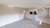 This single-story space is designed with a clean, modern aesthetic, highlighted by light gray laminate flooring and warm neutral-toned walls that carry consistently throughout. The open-concept living area flows directly into the kitchen, which is separated by a peninsula counter finished with granite-style countertops. The kitchen is equipped with white cabinetry, black appliances including a refrigerator and range, and tile flooring, providing a functional contrast to the living space.

The living area benefits from natural light entering through a wide window, with views of the surrounding neighborhood. A wall-mounted HVAC unit provides climate control. Multiple bedrooms are present, each featuring the same laminate flooring, neutral walls, and generously sized windows that invite natural light and outdoor views, including palm trees suggesting a warm-weather locale. One bedroom includes a mirrored sliding-door closet offering practical storage.

The bathroom is finished with tile flooring, a granite-topped vanity with a drop-in sink, a tub-shower combination, and a large wall mirror with overhead lighting. White cabinetry and chrome fixtures complete the clean, functional design. Overall, the space presents a well-maintained, move-in ready environment with consistent finishes throughout.