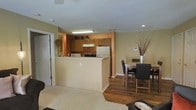 This single-floor residence offers an open-concept layout that seamlessly connects the kitchen, dining area, and living space, creating a functional and cohesive environment. The kitchen is equipped with oak cabinetry providing ample storage, a white refrigerator, a range with overhead ventilation, and a countertop that extends into a peninsula — serving as both a workspace and a casual bar-style seating area. The warm terracotta accent wall in the kitchen adds a distinct pop of color against the neutral beige tones that carry throughout the space. Adjacent to the kitchen, the dining area accommodates a full table and chairs, making it well-suited for everyday meals or entertaining. The transition from the kitchen and dining zone into the living area is marked by a shift in flooring — rich hardwood planks give way to plush carpeting, clearly delineating each functional zone while maintaining an open feel. Recessed lighting and overhead fluorescent fixtures ensure the space is well-lit throughout. A visible thermostat on the wall speaks to the practical, resident-focused design of the home. The overall ambiance is warm, approachable, and move-in ready.