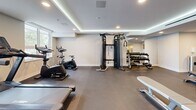 Located at 1110 Lawson Avenue in Kelowna, BC, the Troika fitness centre is a thoughtfully designed exercise space that provides residents with a comprehensive workout facility. The room features a modern, clean aesthetic characterized by a coffered ceiling with elegant LED cove lighting, recessed pot lights, and crisp white walls that create a bright and motivating atmosphere.

The space is divided into functional zones to accommodate a variety of workout styles. The cardio area, positioned along a large window that allows ample natural light, is equipped with Spirit treadmills and stationary cycling bikes, offering an energizing environment for endurance training. Nearby, a multi-functional cable machine and a full rack of dumbbells supported by a mirrored wall cater to strength training needs. Workout benches are available throughout the open floor area, which features durable dark rubber flooring suited for high-impact activity.

A dedicated stretching and floor exercise zone includes exercise mats, while an open shelving unit provides convenient storage. A built-in water station with a bottle filler and a waste bin ensure practicality and comfort during workouts. The Troika branding is prominently displayed on a slatted wood feature wall, adding a polished, boutique-gym feel to this well-appointed community fitness centre.