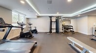 Located at 1110 Lawson Avenue in Kelowna, BC, the Troika fitness centre is a thoughtfully designed exercise space that provides residents with a comprehensive workout facility. The room features a modern, clean aesthetic characterized by a coffered ceiling with elegant LED cove lighting, recessed pot lights, and crisp white walls that create a bright and motivating atmosphere.

The space is divided into functional zones to accommodate a variety of workout styles. The cardio area, positioned along a large window that allows ample natural light, is equipped with Spirit treadmills and stationary cycling bikes, offering an energizing environment for endurance training. Nearby, a multi-functional cable machine and a full rack of dumbbells supported by a mirrored wall cater to strength training needs. Workout benches are available throughout the open floor area, which features durable dark rubber flooring suited for high-impact activity.

A dedicated stretching and floor exercise zone includes exercise mats, while an open shelving unit provides convenient storage. A built-in water station with a bottle filler and a waste bin ensure practicality and comfort during workouts. The Troika branding is prominently displayed on a slatted wood feature wall, adding a polished, boutique-gym feel to this well-appointed community fitness centre.