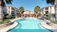 This ground-floor space is part of a well-appointed multi-story residential complex designed with a Mediterranean-inspired aesthetic. The exterior showcases warm stucco façades, decorative stonework, and balconied upper floors, all centered around a standout resort-style pool area. The freeform pool, framed by tall palm trees and a wooden pergola, is surrounded by open patio seating, creating a welcoming communal outdoor environment beneath clear skies.

The covered ground-floor corridors serve as the primary circulation spaces, connecting residents to key building amenities. These passageways feature horizontal lap-siding walls in neutral tones, textured ceilings, and flush-mounted lighting that provides consistent illumination throughout. A resident elevator offers convenient vertical access between floors, positioned alongside a metal-railed stairwell for additional connectivity.

One covered corridor includes a seating area furnished with white slatted benches, offering a comfortable transitional space that opens toward the pool courtyard, allowing natural light and outdoor views to filter through. Additional utility areas, including mechanical equipment alcoves, are thoughtfully integrated along the building perimeter. Overall, the space balances practical residential infrastructure with an inviting, resort-influenced atmosphere.