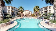 This ground-floor space is part of a well-appointed multi-story residential complex designed with a Mediterranean-inspired aesthetic. The exterior showcases warm stucco façades, decorative stonework, and balconied upper floors, all centered around a standout resort-style pool area. The freeform pool, framed by tall palm trees and a wooden pergola, is surrounded by open patio seating, creating a welcoming communal outdoor environment beneath clear skies.

The covered ground-floor corridors serve as the primary circulation spaces, connecting residents to key building amenities. These passageways feature horizontal lap-siding walls in neutral tones, textured ceilings, and flush-mounted lighting that provides consistent illumination throughout. A resident elevator offers convenient vertical access between floors, positioned alongside a metal-railed stairwell for additional connectivity.

One covered corridor includes a seating area furnished with white slatted benches, offering a comfortable transitional space that opens toward the pool courtyard, allowing natural light and outdoor views to filter through. Additional utility areas, including mechanical equipment alcoves, are thoughtfully integrated along the building perimeter. Overall, the space balances practical residential infrastructure with an inviting, resort-influenced atmosphere.