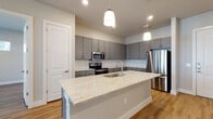 This well-appointed single-floor residence showcases a thoughtfully designed open-concept layout that seamlessly connects the kitchen and living areas. The kitchen serves as the centerpiece of the home, featuring sleek gray shaker-style cabinetry, a classic white subway tile backsplash, and a generously sized island topped with light granite countertops. An undermount sink and stylish pendant lighting add refined detail, while a full suite of stainless steel appliances — including a range, built-in microwave, and refrigerator — ensures modern functionality. Warm hardwood-style flooring flows consistently throughout the main living spaces and bedrooms, contributing to a cohesive and inviting atmosphere. The bedrooms are well-proportioned, receiving natural light through large windows that offer views of the surrounding greenery. Each bedroom benefits from dedicated closet space, with the primary suite featuring a spacious walk-in closet. The primary bathroom is particularly notable, offering a dual-sink vanity with stone countertops, large-format tile flooring, a glass-enclosed walk-in shower adorned with marble-look tile, and a separate soaking tub. A secondary bathroom mirrors the clean, contemporary aesthetic with quality tile work and efficient layout. Built-in shelving and ample storage throughout complete this modern, move-in-ready home.