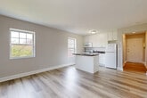 Located at 7112-7312 John F Kennedy Blvd E in North Bergen, NJ, this efficiently laid-out single-floor unit offers a thoughtfully organized living space. Entry is through a narrow hallway finished in warm neutral tones, with light wood-style flooring that continues throughout the unit, lending a cohesive and clean aesthetic.

The open-plan living, dining, and kitchen area forms the heart of the unit. The kitchen is well-appointed with white cabinetry, a dark granite-style countertop peninsula that doubles as a breakfast bar, white appliances including a gas range, microwave, and refrigerator, and a dedicated sink. Multiple windows allow natural light to fill the space, creating a bright and airy atmosphere.

The living and dining area adjoins the kitchen seamlessly, offering a functional and open layout. A cast-iron radiator provides supplemental heating, and additional doors provide access to utility or storage spaces.

The bathroom is finished in a crisp, all-white palette featuring large marble-look floor tiles, white subway wall tiles, a vanity with storage, and a window that allows natural light to brighten the space. The overall design of the unit is clean, modern, and practical, making efficient use of every square foot available.