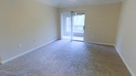 This single-floor space features a generously sized living area finished with neutral-toned wall-to-wall carpeting that lends a warm, comfortable feel underfoot. The room is bathed in natural light courtesy of a large sliding glass door fitted with vertical blinds, which can be adjusted to control light and privacy. The sliding door provides direct access to a private balcony, offering pleasant outdoor views of surrounding greenery. The walls are painted in a soft, neutral tone that complements the light carpeting and contributes to an airy, open ambiance throughout the space. A ceiling light fixture is centrally positioned overhead, providing functional overhead illumination. Clean white baseboards frame the perimeter of the room, adding a crisp, finished look to the interior. The overall design is straightforward and practical, providing a versatile layout suitable for a variety of furniture arrangements. The connection between the indoor living area and the outdoor balcony enhances the sense of space and allows for easy enjoyment of natural surroundings.