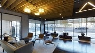 This single-floor space makes a striking architectural statement through its thoughtful combination of natural and industrial materials. The exposed timber truss ceiling adds warmth and organic texture overhead, while sleek dark structural elements and polished dark-tile flooring ground the space in a refined, modern aesthetic. Ambient drum pendant lighting suspended from the ceiling provides a soft, welcoming glow that complements the abundant natural light streaming in through expansive floor-to-ceiling windows. These large-pane windows wrap along the exterior-facing walls, offering unobstructed views of the surrounding streetscape and flooding the interior with daylight. The open layout is furnished with a curated selection of contemporary seating, including low-profile leather lounge chairs with slim metal frames and plush modular sofas, arranged thoughtfully around area rugs and accent tables to define intimate gathering areas. The overall ambiance strikes a balance between industrial character and modern sophistication, making the space feel both dynamic and comfortable. The transparency of the glazed facade creates a strong visual connection between the interior and the outdoor environment, enhancing the sense of openness and scale throughout.