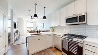 This single-floor residence is designed with a clean, contemporary aesthetic that balances functionality and style. The open-concept kitchen serves as a central hub, featuring white shaker cabinetry extending to the ceiling, a white quartz peninsula with an undermount sink and seating for multiple bar stools, and a white subway tile backsplash. Stainless steel appliances — including a gas range, over-the-range microwave, dishwasher, and French door refrigerator — complete the well-equipped space. Two matte black pendant lights hang above the peninsula, adding a modern accent. Warm-toned hardwood flooring flows consistently throughout the unit, unifying each room.

The living area is bright and inviting, benefiting from a large window that draws in natural light. A ceiling fan provides comfort, and the space connects to a compact dining nook. Both bedrooms are comfortably proportioned, each with windows that allow natural light; one features an exterior view with what appears to be a balcony railing visible beyond. The bathroom is well-appointed with a wide white vanity, large framed mirror, glass-enclosed walk-in shower with tiled walls, and wood-look flooring, offering a spa-like finish consistent with the unit's overall modern sensibility.