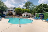 Located at 300 Cityline Ave NE in Atlanta, Georgia, this outdoor amenity space at Muse ATL offers a resort-style pool experience designed for relaxation and social gathering. The centerpiece is a generously sized, freeform swimming pool with crystal-clear turquoise water, providing ample room for both leisure and recreation. The pool's curvilinear design adds a dynamic visual appeal to the space.

Adjacent to the pool, a covered open-air cabana serves as a social hub, featuring a sloped roof supported by clean white columns. Inside the cabana, high-top bar seating is arranged along a built-in counter, accompanied by mounted televisions and an outdoor kitchen or grilling station — ideal for entertaining and gatherings.

The surrounding pool deck is finished in light-toned concrete and is outfitted with blue lounge chairs and patio umbrellas, creating a comfortable sunbathing environment. Mature trees frame the perimeter of the space, providing natural shade and a sense of privacy while softening the urban setting.

Decorative landscaping elements, including potted plants and lush greenery along the fence line, enhance the atmosphere. The Muse ATL branded signage, displayed prominently near a living green wall feature, adds a distinct identity to the space. Multi-story residential buildings visible in the background contextualize this amenity within a vibrant Atlanta community.