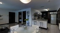 This single-floor space showcases a contemporary design aesthetic defined by a striking black-and-white color palette. The open-plan layout seamlessly connects the dining and kitchen areas, creating a cohesive and functional living environment.

The dining area features a round glass-top table paired with modern white chairs, adding a clean, minimalist touch. A decorative glass vase with a green plant serves as a centerful centerpiece, introducing a natural accent against the otherwise monochromatic scheme.

The kitchen is well-appointed with dark countertops, a raised breakfast bar with chrome bar stools, and upper white cabinetry. Appliances and fixtures are neatly integrated into the design, while a decorative black-framed artwork on the kitchen wall adds a sophisticated visual element.

A partial wall displays a vertical series of abstract black-and-white artworks, serving as both a design feature and a subtle divider between zones. Dark-framed doors lead to additional rooms, one of which reveals a glimpse of a bedroom. Overhead, a large spherical ceiling light provides soft, ambient illumination throughout the space, complementing the modern and polished overall ambiance.