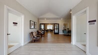 This single-story community clubhouse is designed to offer a well-rounded amenity experience with a thoughtful, open layout. The entry foyer makes an immediate impression with its vaulted ceiling, crown molding, and light wood-style flooring that extends throughout much of the facility. Framed artwork and comfortable seating areas flank the entryway, creating a welcoming first impression.

The main lounge area is generously proportioned, featuring multiple seating arrangements including teal upholstered sofas, leather accent chairs, and a sliding barn door detail that adds a modern farmhouse character. Adjacent to the lounge, a well-equipped community kitchen features dark espresso cabinetry, granite countertops, a large island with bar seating, pendant lighting, and stainless steel appliances, including a full-size refrigerator. A built-in desk area provides additional workspace.

A sun-filled dining nook with large windows and a round table offers a more intimate gathering spot. A dedicated fitness room is outfitted with cardio machines, a full dumbbell rack, medicine balls, and resistance equipment, with natural light provided by exterior-facing windows. The facility also includes a shared laundry room lined with commercial-grade washers and dryers. Neutral wall tones, crown molding, and recessed lighting unify the space throughout.