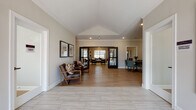 This single-story community clubhouse is designed to offer a well-rounded amenity experience with a thoughtful, open layout. The entry foyer makes an immediate impression with its vaulted ceiling, crown molding, and light wood-style flooring that extends throughout much of the facility. Framed artwork and comfortable seating areas flank the entryway, creating a welcoming first impression.

The main lounge area is generously proportioned, featuring multiple seating arrangements including teal upholstered sofas, leather accent chairs, and a sliding barn door detail that adds a modern farmhouse character. Adjacent to the lounge, a well-equipped community kitchen features dark espresso cabinetry, granite countertops, a large island with bar seating, pendant lighting, and stainless steel appliances, including a full-size refrigerator. A built-in desk area provides additional workspace.

A sun-filled dining nook with large windows and a round table offers a more intimate gathering spot. A dedicated fitness room is outfitted with cardio machines, a full dumbbell rack, medicine balls, and resistance equipment, with natural light provided by exterior-facing windows. The facility also includes a shared laundry room lined with commercial-grade washers and dryers. Neutral wall tones, crown molding, and recessed lighting unify the space throughout.