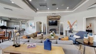 This single-floor community clubhouse is designed to serve as a sophisticated social hub, combining comfortable gathering spaces with upscale amenities. The main lounge area is an expansive, open-concept room defined by multi-layered tray ceilings accented in slate gray, recessed lighting, and a striking sculptural chandelier with interlocking bronze rings. A floor-to-ceiling stacked stone fireplace serves as the room's focal point, flanked by decorative vases and topped with a wall-mounted television. Generously sized sectional sofas in neutral gray tones are arranged throughout, accented with navy, rust, and gold throw pillows that complement the contemporary artwork adorning the walls. Light-toned tile flooring runs throughout the space, enhancing the bright, airy atmosphere. Large windows and glass-paned French doors flood the interior with natural light while offering views of the surrounding outdoor areas. A dedicated café-style corner features a Starbucks coffee station with granite countertops, bar seating, white cabinetry, and stainless steel appliances. Adjacent to this area is a casual dining nook with a round table and upholstered chairs, positioned beside tall windows overlooking the exterior. Arched doorways and clean architectural lines connect each zone seamlessly, creating a cohesive and welcoming environment throughout.
