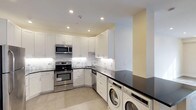 This single-floor space showcases a well-appointed kitchen designed with both functionality and style in mind. The kitchen features floor-to-ceiling white shaker-style cabinetry that maximizes storage, complemented by sleek dark countertops that provide a striking contrast. The marble-patterned backsplash adds a refined touch, running the length of the cooking and prep areas.

Stainless steel appliances, including a gas range, built-in microwave, and dishwasher, are seamlessly integrated into the cabinetry, giving the kitchen a cohesive and polished look. Recessed lighting throughout the ceiling ensures bright, even illumination across the workspace.

A notable feature of this space is the inclusion of an in-unit washer and dryer, conveniently positioned beneath an extension of the dark countertop, providing additional surface area while keeping laundry appliances neatly tucked away.

Light-toned tile flooring runs throughout, contributing to an open and clean aesthetic. The overall design balances practicality with a contemporary style, making efficient use of the available space while maintaining a modern, streamlined appearance.
