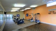 This single-floor fitness center offers a practical and well-organized space for exercise and wellness. The room is defined by a vibrant orange accent wall that energizes the environment, complemented by neutral-toned walls and a combination of carpeted and hardwood-style flooring that divides the space into distinct workout zones. Overhead, multiple flush-mounted panel lights and ceiling fans provide consistent, comfortable illumination and airflow throughout the room. The cardio zone is outfitted with several machines, including an elliptical trainer and a recumbent stationary bike, ideal for low-impact aerobic workouts. A multi-station cable and weight machine anchors the strength training area, offering a variety of resistance exercises in a compact, integrated unit. Motivational fitness artwork is displayed along the walls, adding character and purpose to the space. Ventilation is supported by visible ceiling vents, ensuring a comfortable climate during use. The layout is thoughtfully arranged to allow ease of movement between equipment, making the space functional for individual users across all fitness levels.